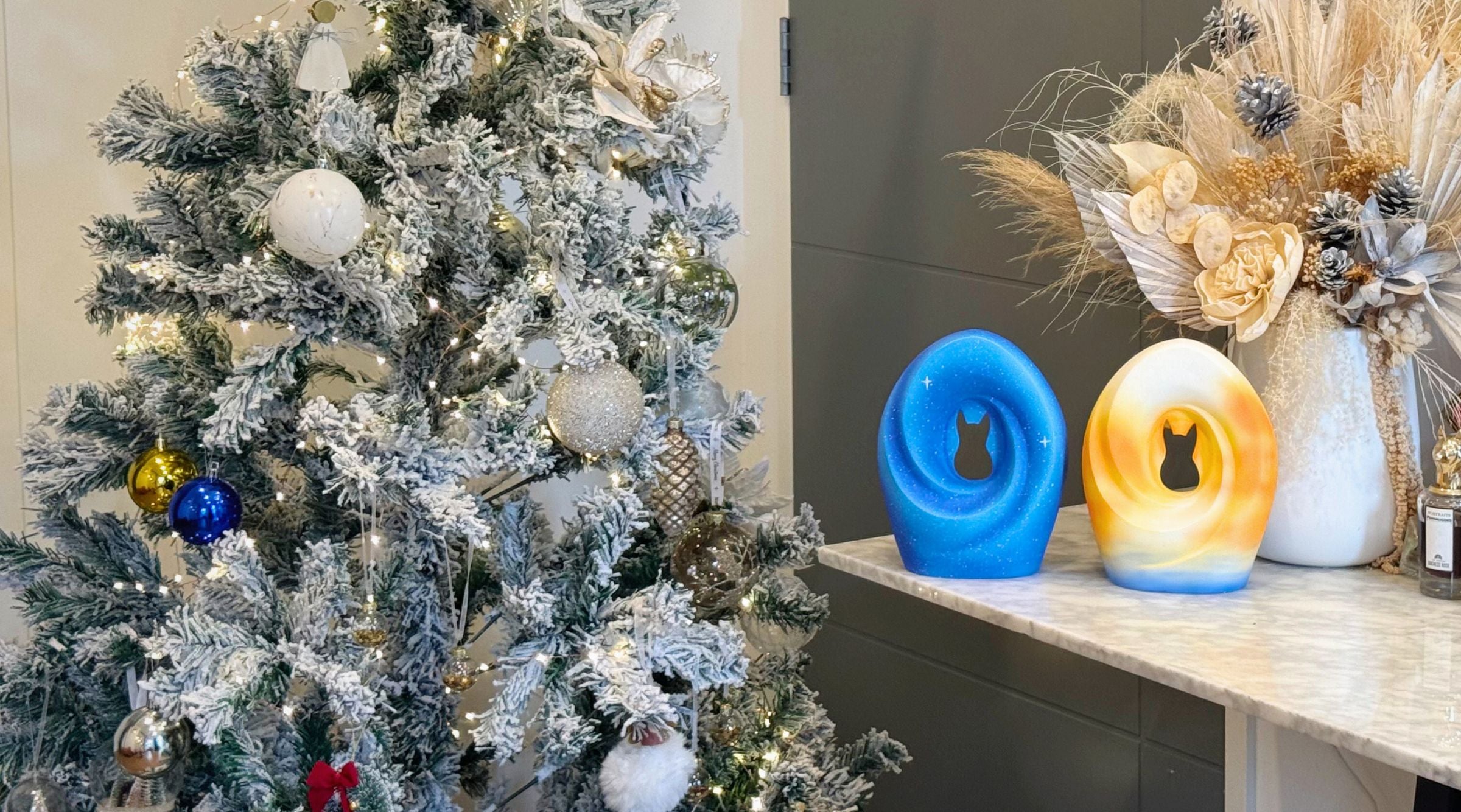 A festive display featuring two pet urns—one in blue with starry details and another in golden hues, each shaped with a silhouette of a cat or dog—placed on a marble countertop next to a vase of dried flowers and a Christmas tree adorned with silver and gold ornaments. The scene radiates warmth, love, and connection.