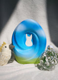 Load image into Gallery viewer, A blue and green gradient pet urn with a carved silhouette, resembling soft clouds and a peaceful meadow. The urn is placed on a light surface alongside a plush bird and a clear glass ornament filled with green leaves and flowers.
