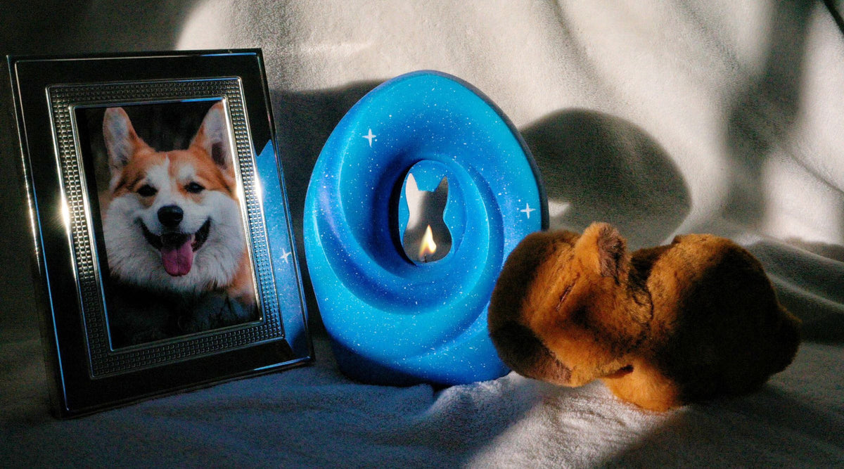 A blue donut-shaped pet urn with a glowing candle silhouette, a framed photo of a happy corgi, and a soft toy on a cozy background, creating a warm and commemorative pet memorial setup.