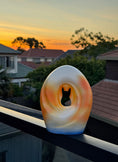 Load image into Gallery viewer, The Sunset Memories Pet Urn featuring a modern cat and dog silhouette carved into an abstract crescent moon design. The urn has warm gradient tones of orange and yellow, resembling a sunset, set against a backdrop of a serene evening sky.
