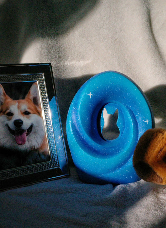 Handcrafted pet urn with a celestial blue design featuring a cut-out silhouette of a cat, adorned with subtle star patterns, placed next to a framed photo of a happy corgi and a plush toy on a soft fabric background under gentle lighting.