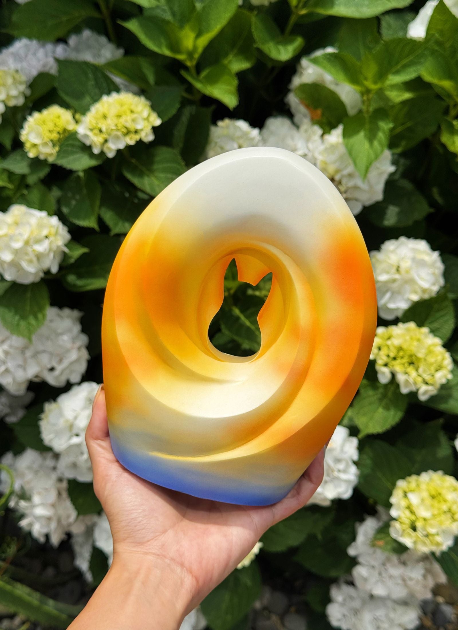Handcrafted pet urn with a sunset gradient design and a cut-out cat silhouette, held in hand against a vibrant garden backdrop filled with blooming white and yellow hydrangeas.
