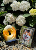 Load image into Gallery viewer, Handcrafted pet urn with a sunset gradient design and a cut-out silhouette of a cat, displayed outdoors among white hydrangeas and greenery, placed next to a framed photo of a smiling corgi on a bed of small pebbles.
