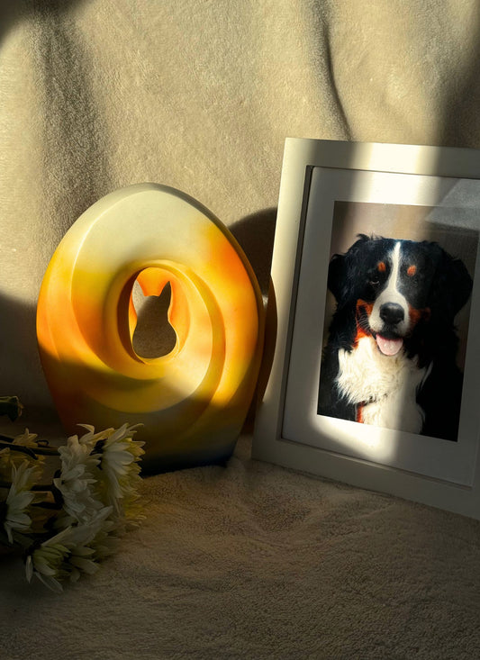Handcrafted pet urn with a unique sunset gradient design, featuring a cut-out silhouette of a cat, placed next to a framed photo of a happy dog and white flowers on a soft fabric background, bathed in warm natural light.